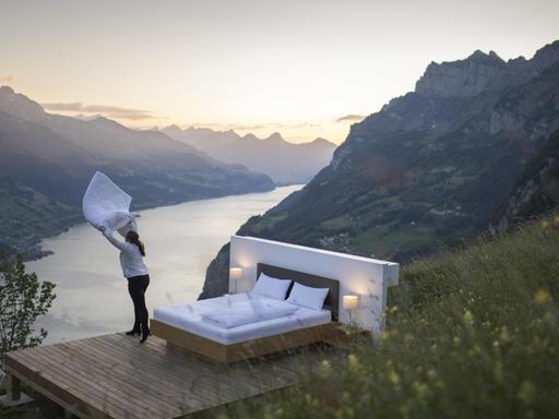 Ein Bett steht inmitten von Bergen, die durch einen Fluss getrennt sind und eine Frau schüttelt die Bettdecke auf. Es ist eine Kollaboration der Schweizer Künstler Frank und Patrik Riklin und der Tourismusbranche.