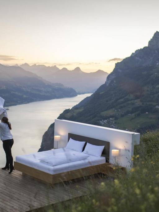 Ein Bett steht inmitten von Bergen, die durch einen Fluss getrennt sind und eine Frau schüttelt die Bettdecke auf. Es ist eine Kollaboration der Schweizer Künstler Frank und Patrik Riklin und der Tourismusbranche.