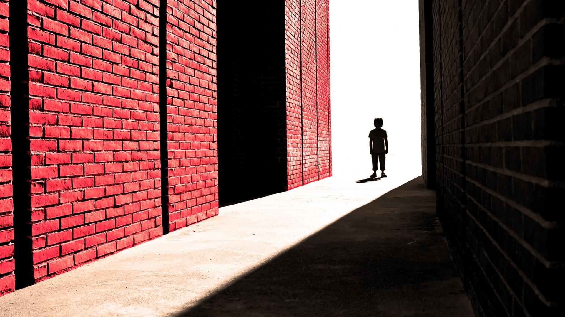 Ein Junge steht weit entfernt in einer Gasse zwischen einem großen Schatten und einer Backsteinmauer.