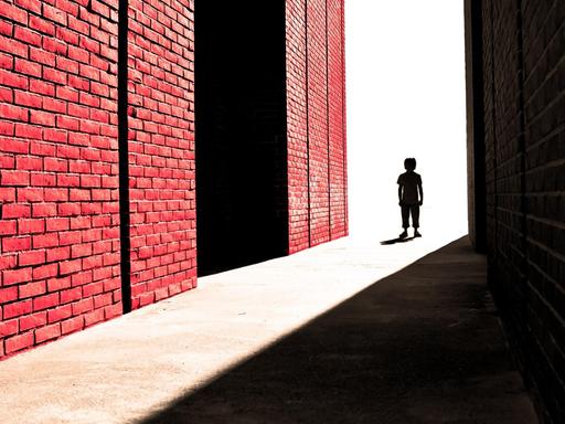 Ein Junge steht weit entfernt in einer Gasse zwischen einem großen Schatten und einer Backsteinmauer.