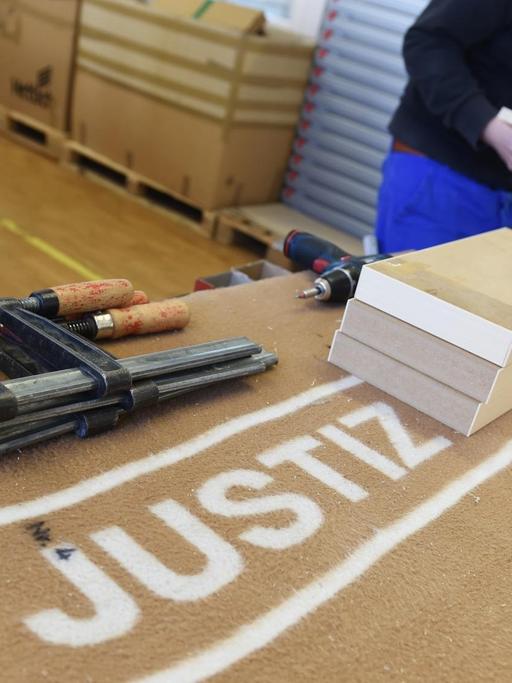 Ein Häftling baut in der Schreinerei der Justizvollzugsanstalt in Ulm (Baden-Württemberg) einen Schrank zusammen, während im Vordergrund mehrere Werkzeuge auf einer Decke liegen, auf der "Justiz" steht.