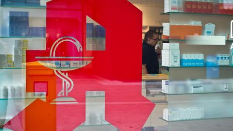 Das rote Apotheken-Logo an einer Eingangstür, dahinter Regale mit Medikamenten.