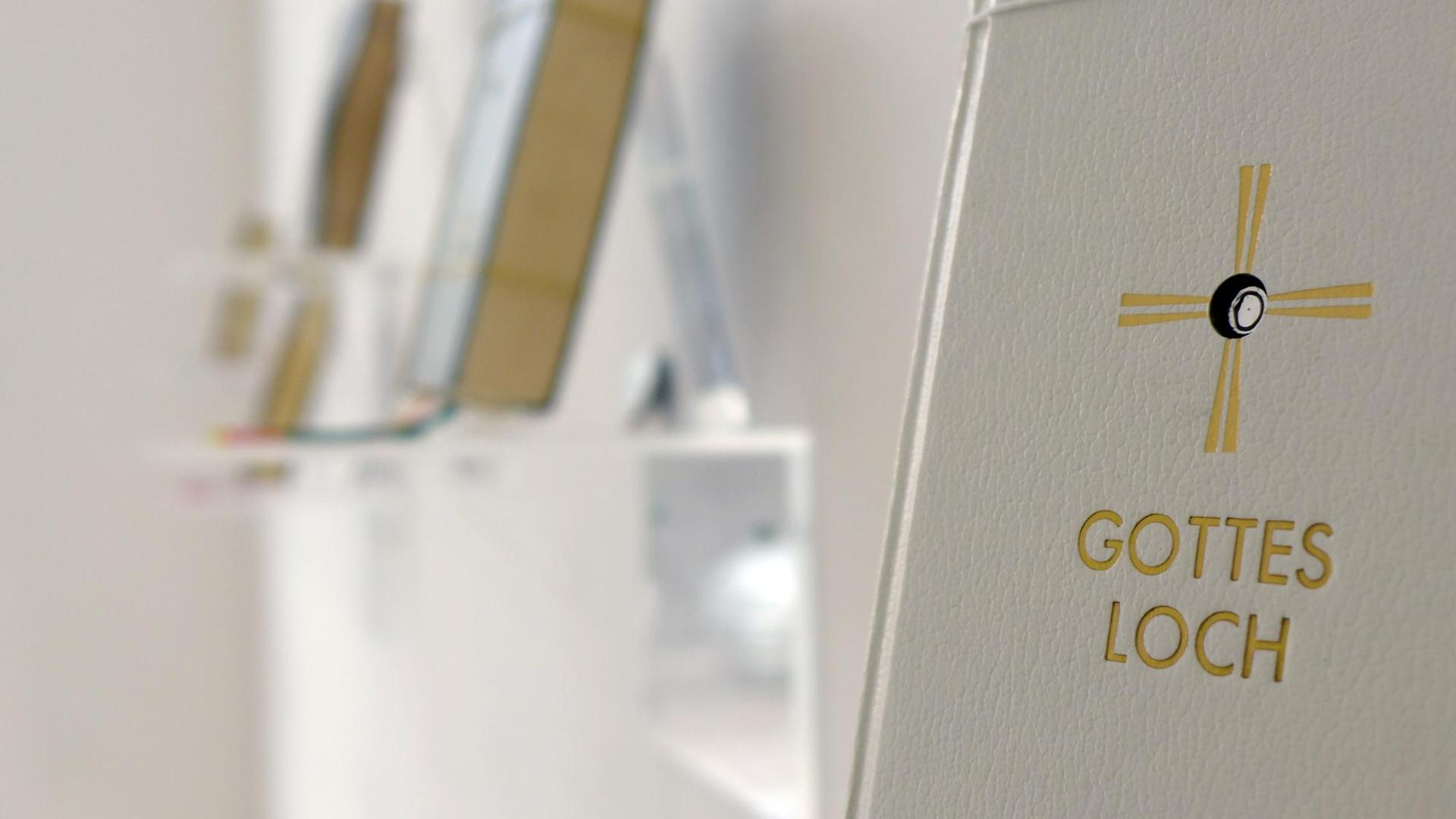 Schrägansicht auf die Installation "Gottesloch" von Georgia Krawiec. Zu sehen ist ein hellgraues Liederbuch mit einem goldenen Kreuz auf dem Cover, in dessen Mitte ein Loch ist. Darunter der Titel "Gottesloch".
