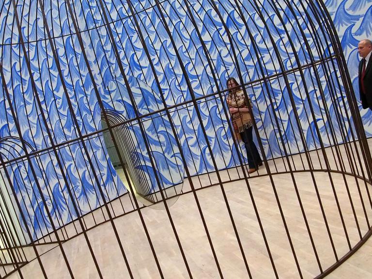 Eine Besucherin und ein Museumsmitarbeiter stehen am 11.11.2015 in der Kunstausstellung "Jeppe Hein. This Way" im Kunstmuseum Wolfsburg vor einem riesigen Käfig, in dem ein Spiegel kreist.