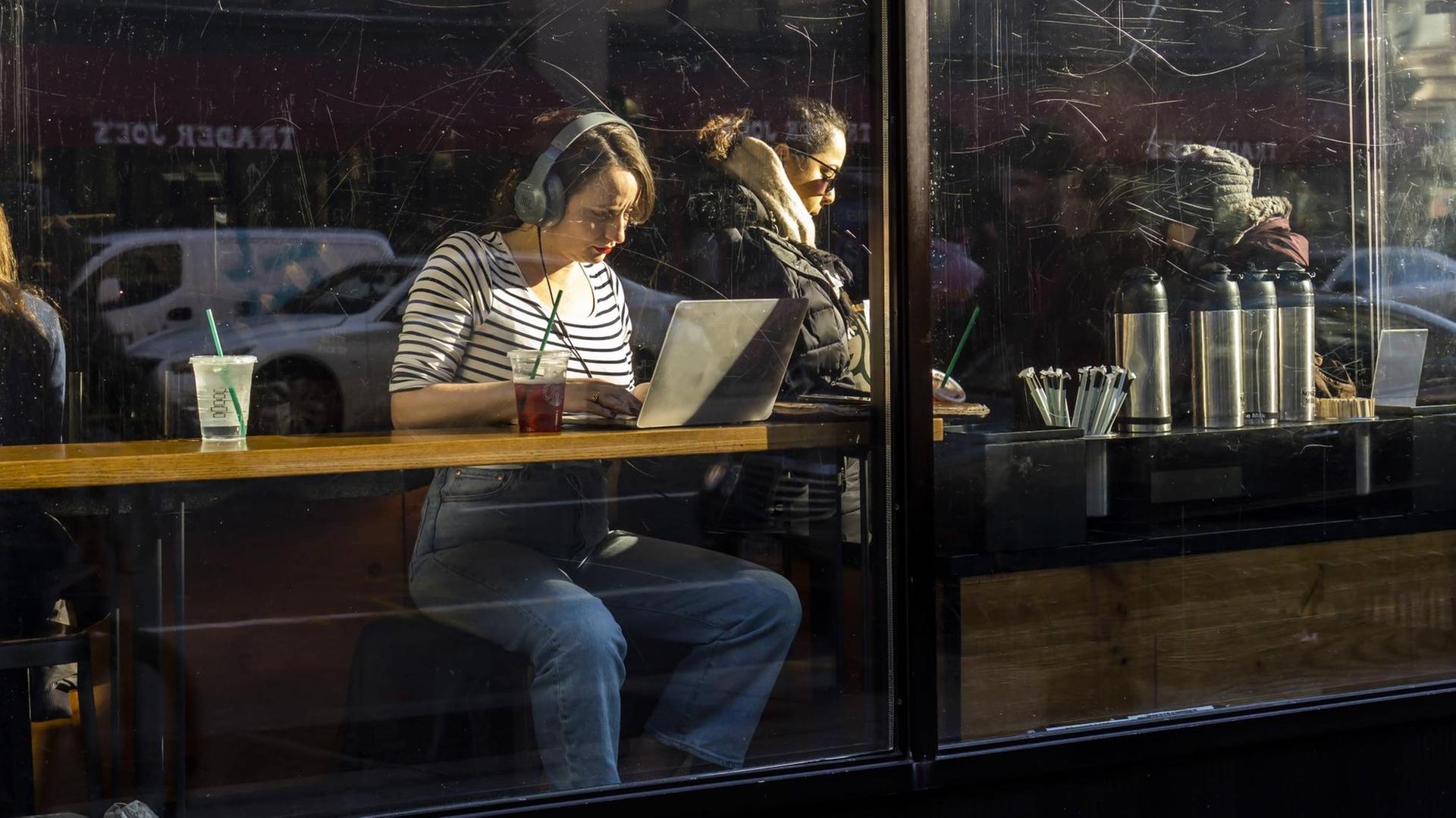Eine Frau sitzt mit Laptop und Kopfhörern am Fenster eines Cafés. Eine Frau sitzt mit Laptop und Kopfhörern am Fenster eines Cafés.