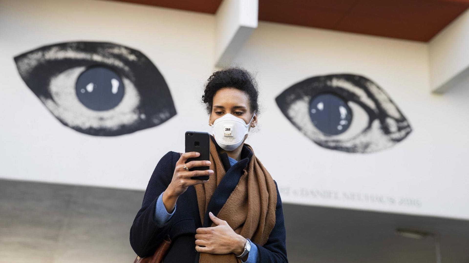 Eine Junge Frau schaut auf ihr Handy mit den Augen eines Grafitti im Hintergrund.