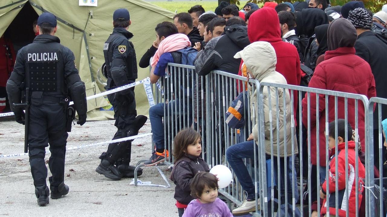 Viele Flüchtlinge in warmen Jacken stehen hinter Absperrgittern. Davor spielen zwei kleine Kinder mit weißen Luftballons. Im Hintergrund sieht man zwei slowenische Polizisten vor einem beigen Zelt.