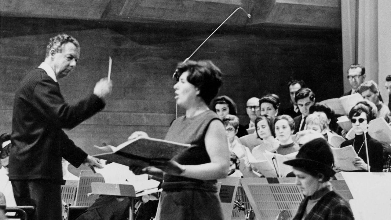 Auf einem schwarz weissen Foto sieht man den Komponisten Benjamin Britten bei der Probe mit dem English Chamber Orchestra und zwei Solistinnen, Miss Heather Harper und Mis Josephine Veasey (sitzend).