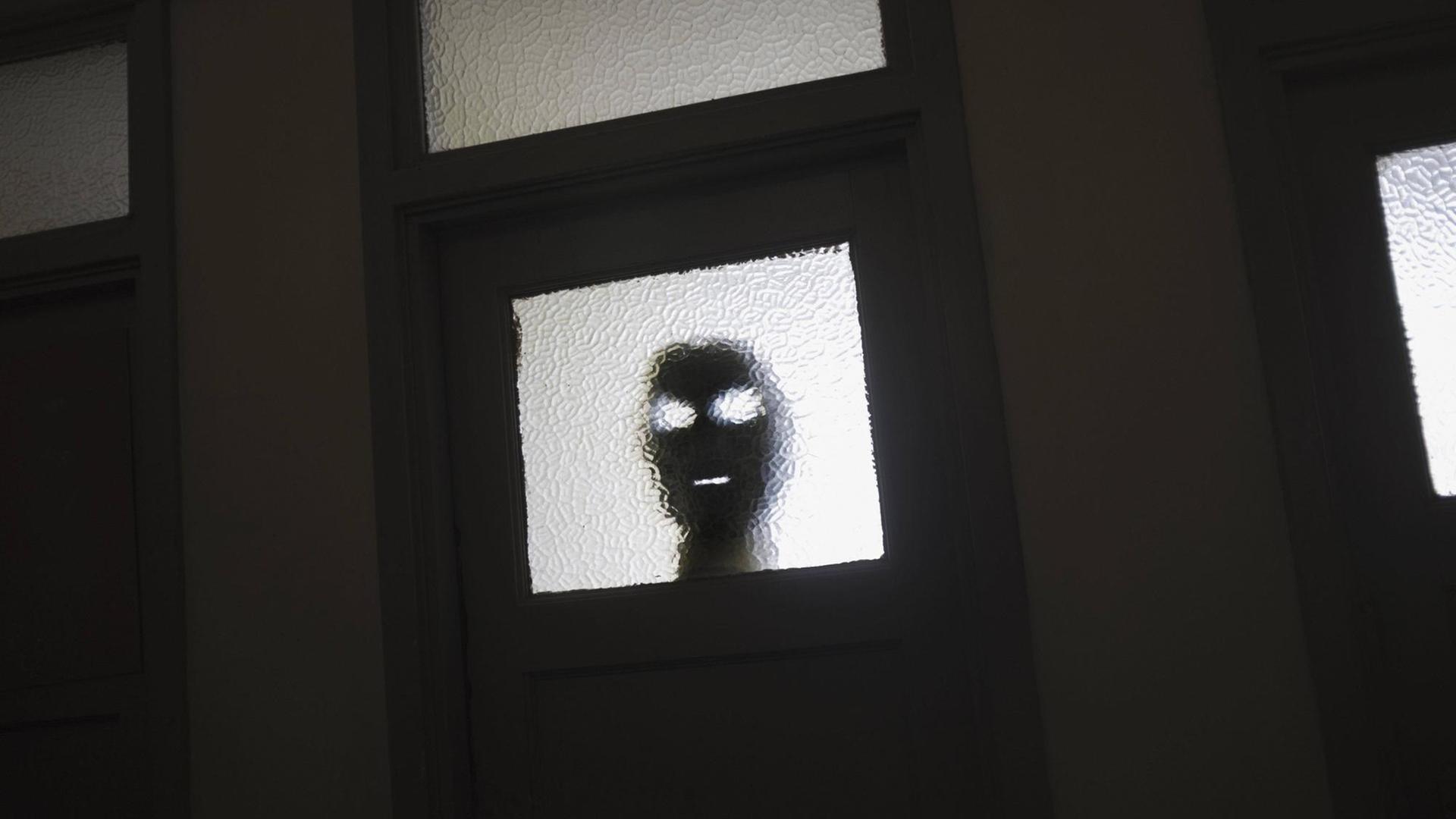 Eine dunkle Maske lauert hinter einem Türfenster.