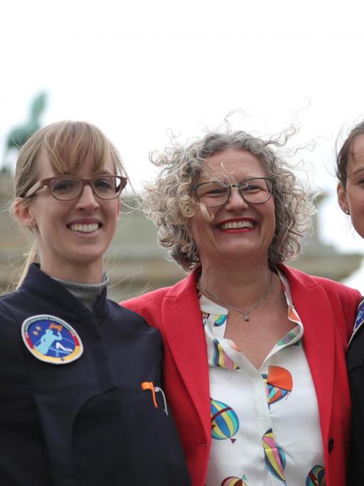 Das Foto zeigt Insa Thiele-Eich und Nicola Baumann, die auf einen Flug ins All zur ISS vorbereitet werden. Zusammen mit der Diplom-Ingenieurin Claudia Kessler stehen sie vor dem Brandenburger Tor.