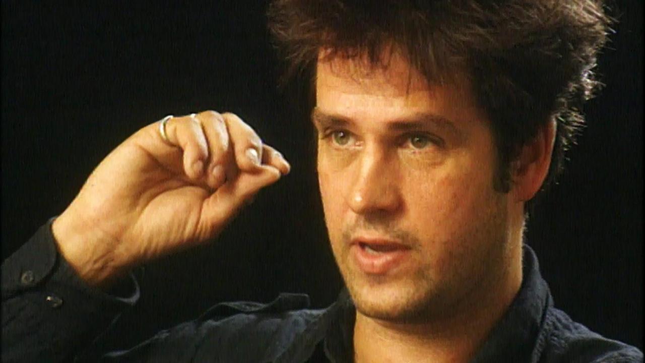 Portrait von Christoph Schlingensief beim Reden vor schwarzem Hintergrund. Er trägt ein schwarzes Hemd und unterstreicht das Gesagte mit einer Geste seiner rechten Hand. Portrait von Christoph Schlingensief beim Reden vor schwarzem Hintergrund. Er trägt ein schwarzes Hemd und unterstreicht das Gesagte mit einer Geste seiner rechten Hand.