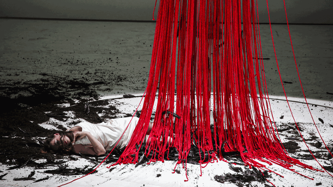 Eine Frau liegt in einem Lichtkegel auf dem Boden, der mit schwarzer Erde bedeckt ist, zudem fallen rote Bänder wie Blut auf sie herab.