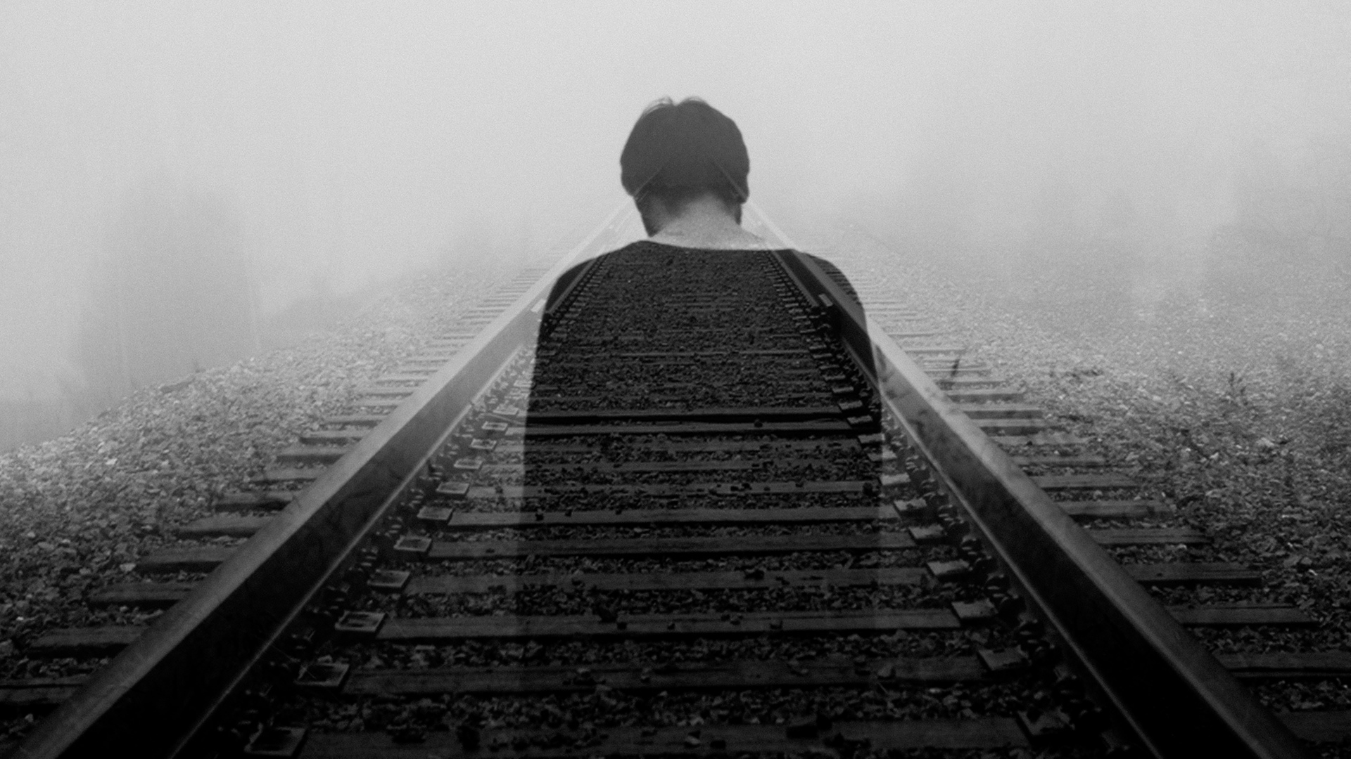 Ein Mann steht mit dem Rücken zur Kamera auf einem Gleisbett und hat den Kopf gesenkt. Im Hintergrund ist es nebelig. Das Foto ist in schwarz-weiß.