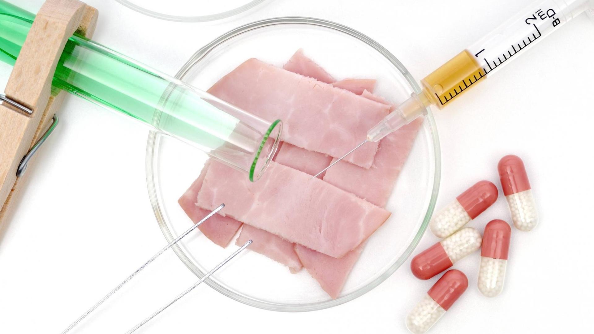 Symbolfoto für Fleisch aus dem Labor - eine Petrischale, ein Reagenzglas und Pillen liegen neben mehreren Scheiben Schinken
