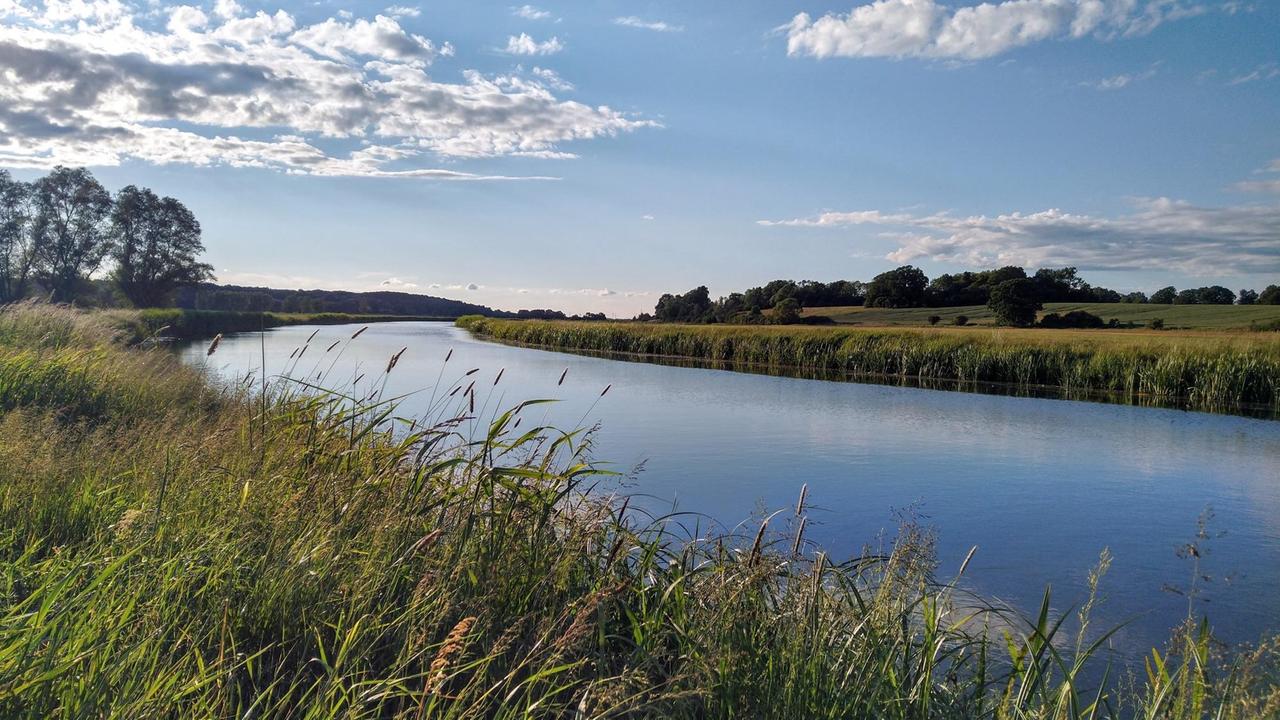 Idyllische Landschaftsaufnahme: Ein tiefblauer Fluss schlängelt sich durch saftige grüne Wiesen, darüber blauer Himmer mit malerischen Wolkenformationen. Idyllische Landschaftsaufnahme: Ein tiefblauer Fluss schlängelt sich durch saftige grüne Wiesen, darüber blauer Himmer mit malerischen Wolkenformationen.