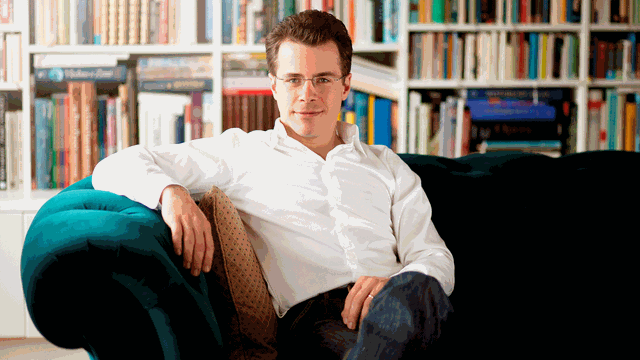 Der Dirigent Jakub Hrůša sitzt auf einem grünen Sofa umgeben von Regalen voller Noten und Bücher.