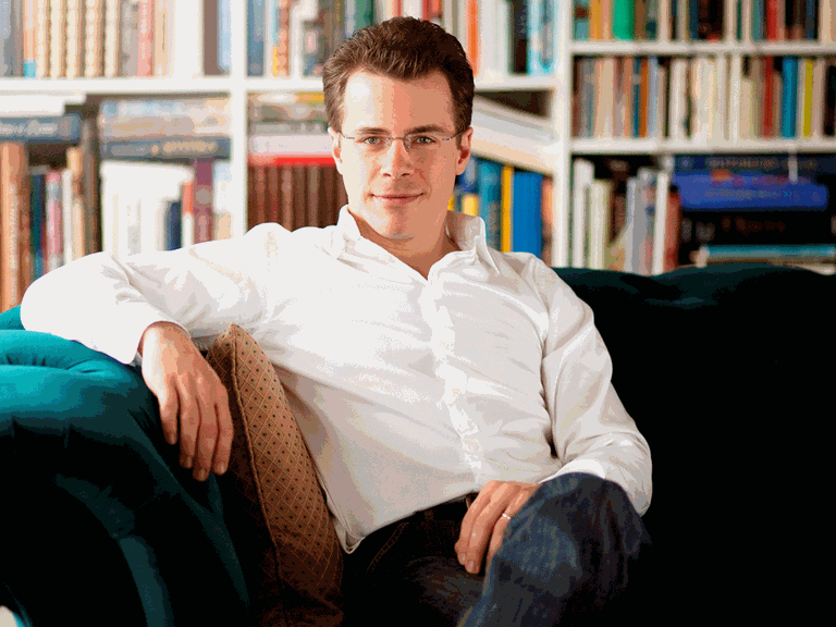 Der Dirigent Jakub Hrůša sitzt auf einem grünen Sofa umgeben von Regalen voller Noten und Bücher.