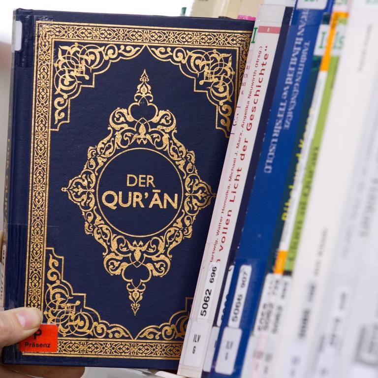 Eine Hand greift in einem Bücherregal einer Bibliothek nach einem Koran.