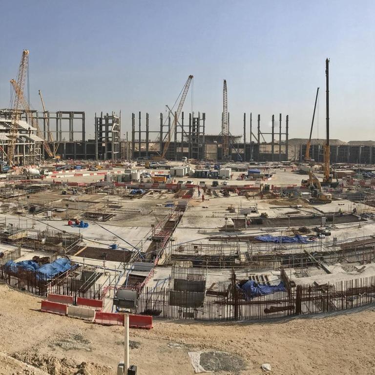 Panoramablick auf die Baustelle eines in Grundzügen bereits erkennbaren Stadions mitten in der Wüste.