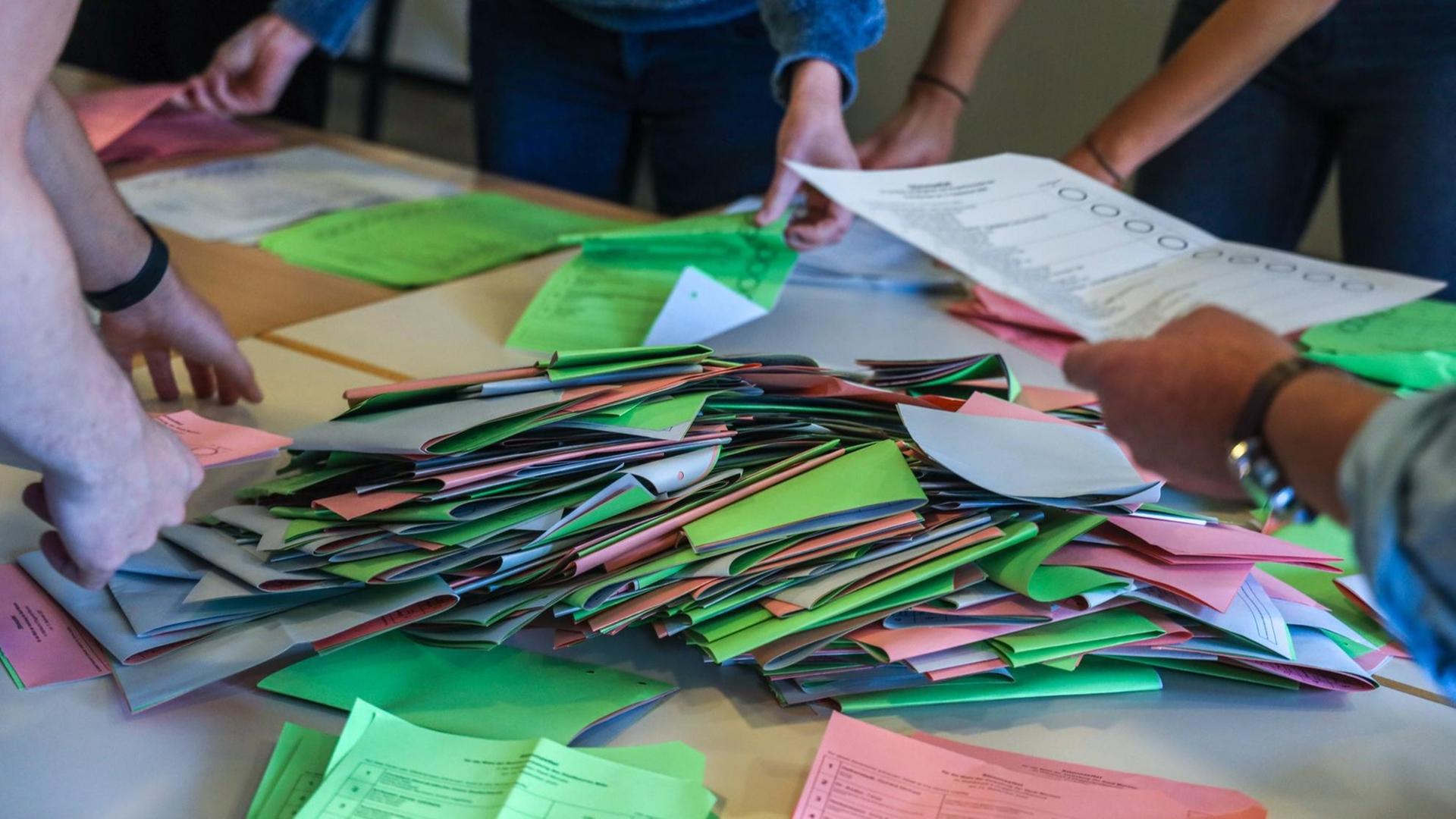 Viele Wahlzettel liegen in Stapeln auf einem Tisch, vier Menschen sortieren sie. Viele Wahlzettel liegen in Stapeln auf einem Tisch, vier Menschen sortieren sie.