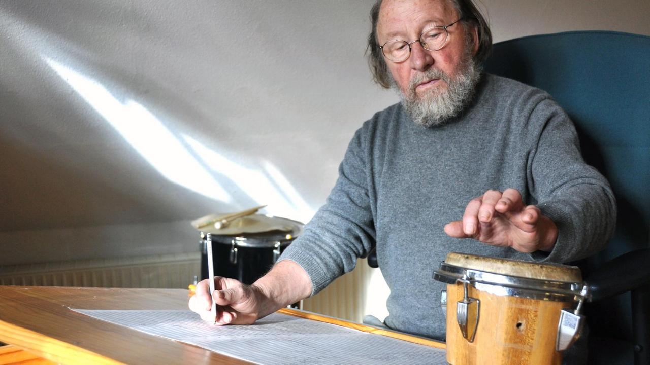 Der Komponist sitzt an einem Schreibtisch mit Notenblatt und kleiner Trommel, die er antippt. Der Komponist sitzt an einem Schreibtisch mit Notenblatt und kleiner Trommel, die er antippt.