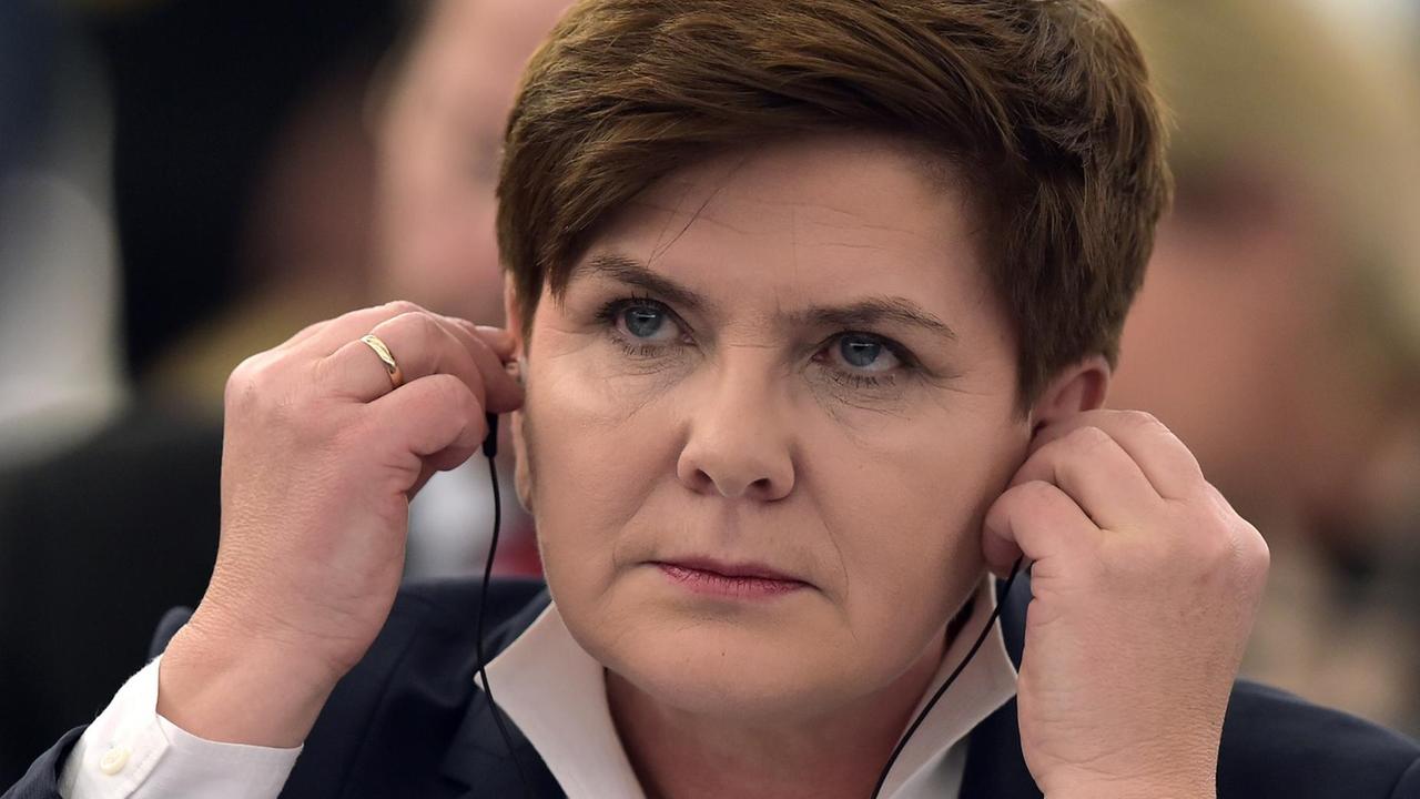 Szydlo mit schwarzem Jackett und weißem Hemd steckt sich gerade die Ohrenstöpsel für den Dolmetscher-Ton in die Ohren. Sie blickt ernst und konzentriert. Szydlo mit schwarzem Jackett und weißem Hemd steckt sich gerade die Ohrenstöpsel für den Dolmetscher-Ton in die Ohren. Sie blickt ernst und konzentriert.