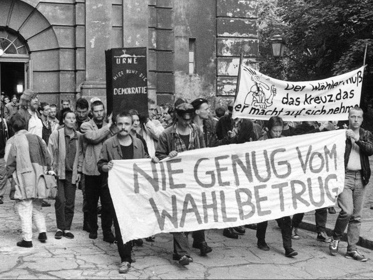 Eine Gruppe von Menschen steht vor einer Kirche und trägt Banner, im Vordergrund eines mit der Aufschrift "Nie genug vom Wahlbetrug".
