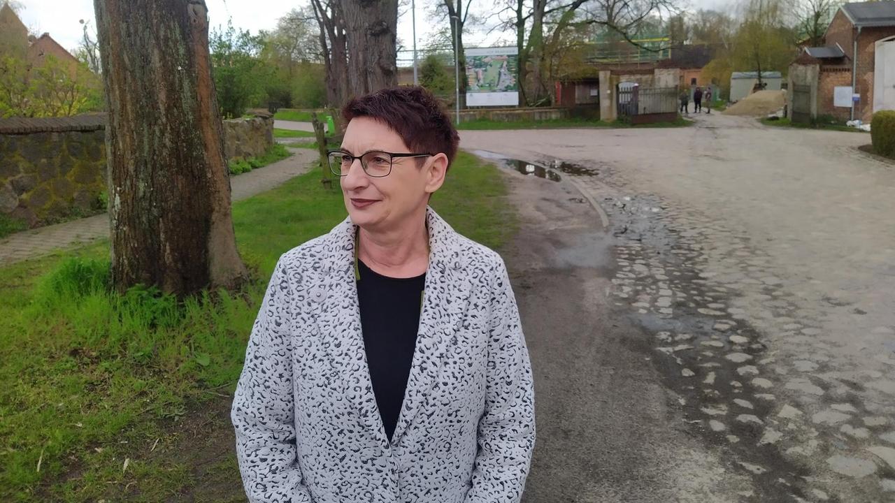 Bürgermeisterin Simone Koß steht auf einer Dorfstraße, links verläuft eine Mauer und es stehen dickstämmige Bäume, rechts ist die Dorfstraße zu sehen. Bürgermeisterin Simone Koß steht auf einer Dorfstraße, links verläuft eine Mauer und es stehen dickstämmige Bäume, rechts ist die Dorfstraße zu sehen.