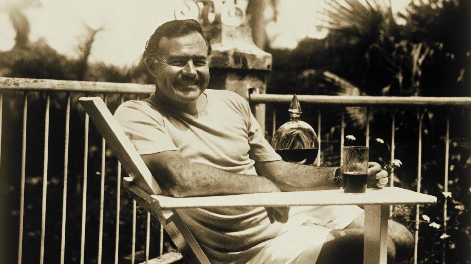Hemingway sitzt mit einem Drink auf der Terrasse und lächelt in die Kamera.