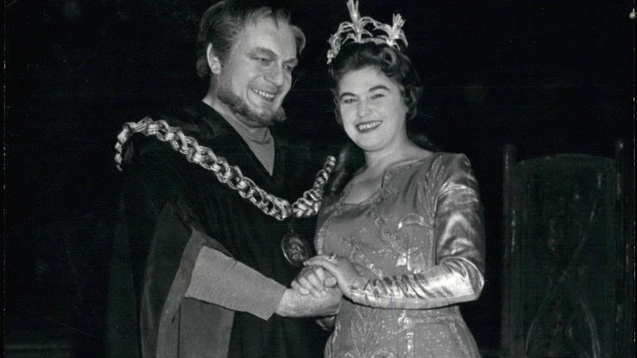 Schwarz-weiß-Foto mit einem Mann (links) und einer Frau: beide lächeln, tragen Kostüme und halten sich an den Händen. Sie trägt eine Krone auf dem Kopf. Schwarz-weiß-Foto mit einem Mann (links) und einer Frau: beide lächeln, tragen Kostüme und halten sich an den Händen. Sie trägt eine Krone auf dem Kopf.