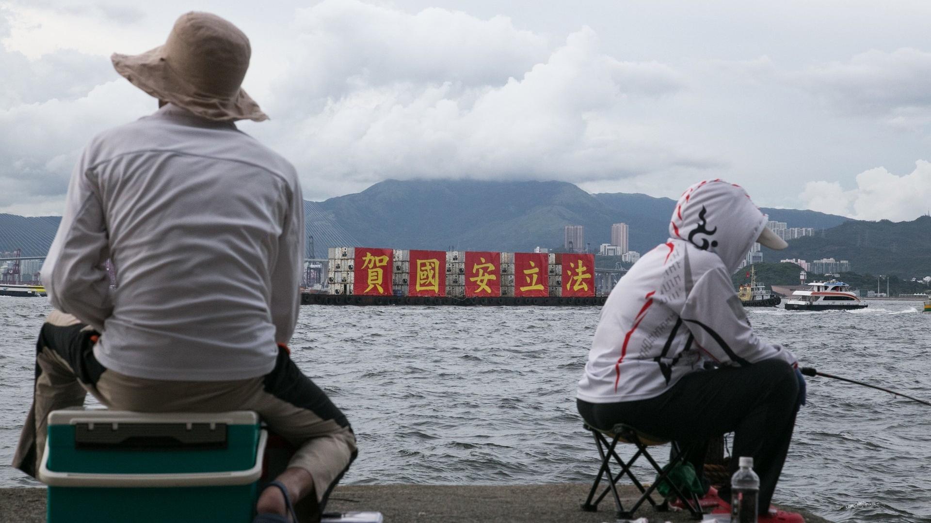 Angler in Hongkong betrachten ein vorüber fahrendes Schiff, welches auf roten Transparenten das von China erlassene neue Sicherheitsgesetz als nationalen Erfolg feiert. Angler in Hongkong betrachten ein vorüber fahrendes Schiff, welches auf roten Transparenten das von China erlassene neue Sicherheitsgesetz als nationalen Erfolg feiert.