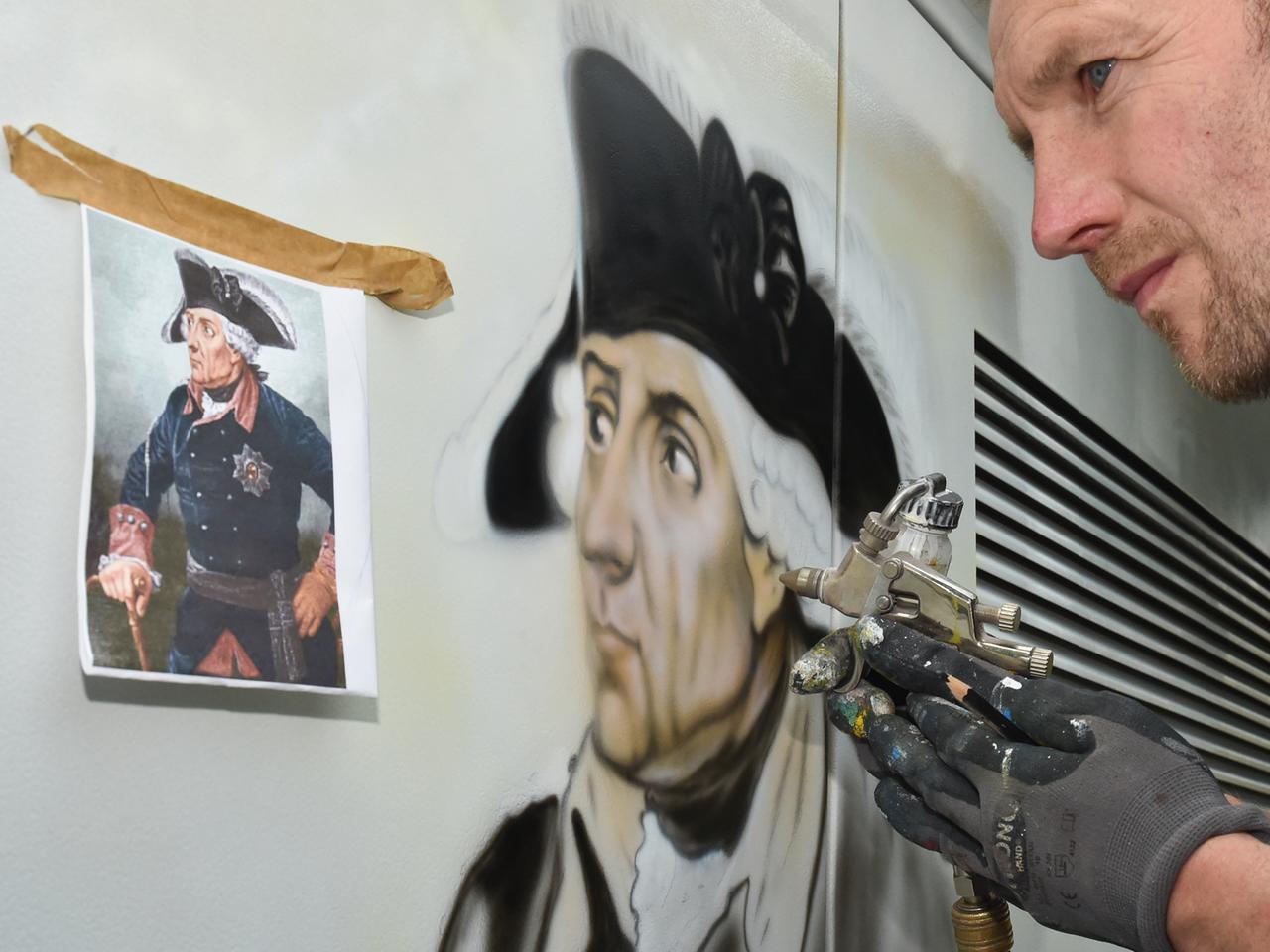 Ein Airbrushmaler sprüht das Porträt von Friedrich II., auch Friedrich der Große oder der Alte Fritz genannt, auf ein Trafohäuschen vor dem Schloss Doberlug in Doberlug-Kirchhain. Ein Airbrushmaler sprüht das Porträt von Friedrich II., auch Friedrich der Große oder der Alte Fritz genannt, auf ein Trafohäuschen vor dem Schloss Doberlug in Doberlug-Kirchhain.