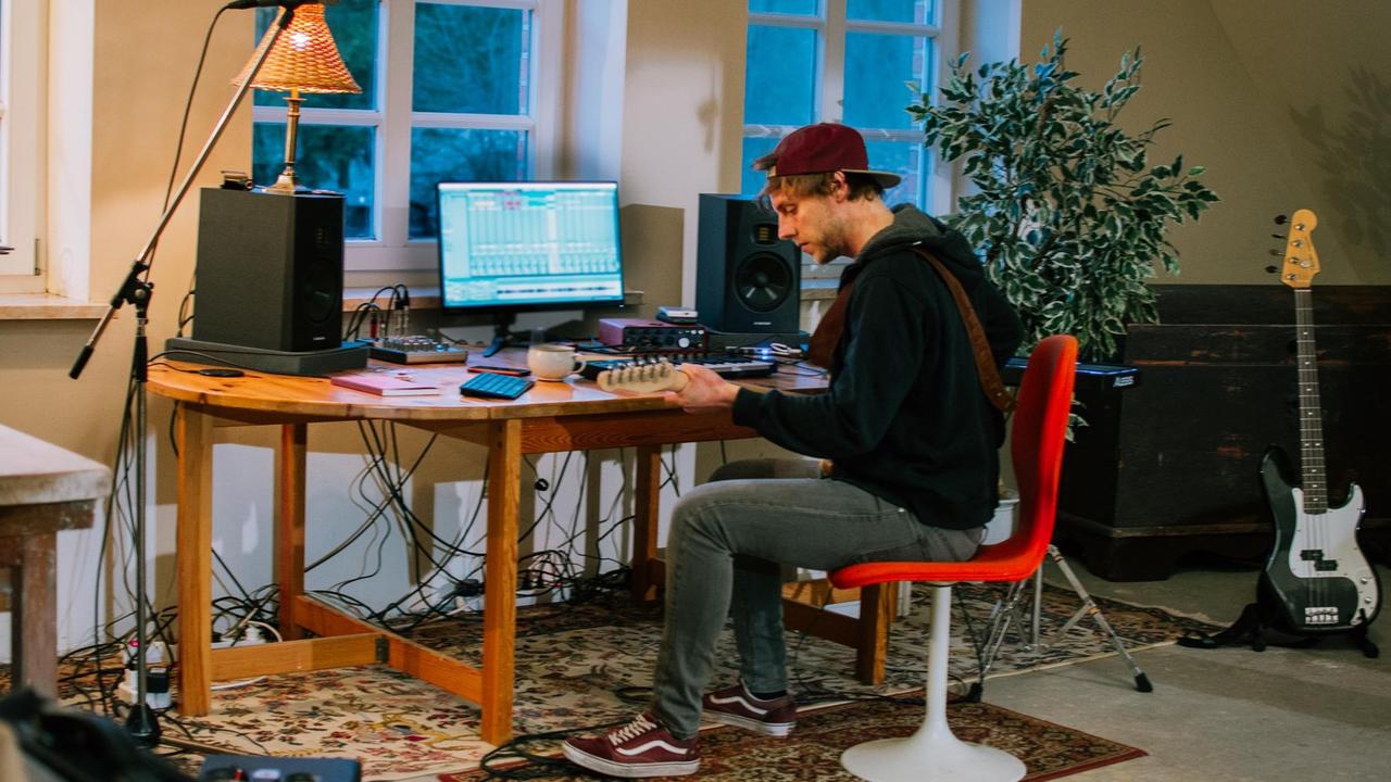 Ein junger Mann mit Baseballkappe sitzt am Schreibtisch vor einem Computer, spielt elektrische Gitarre, die er digital aufnimmt. Rechts neben ihm E-Bass und Zimmerpflanze.