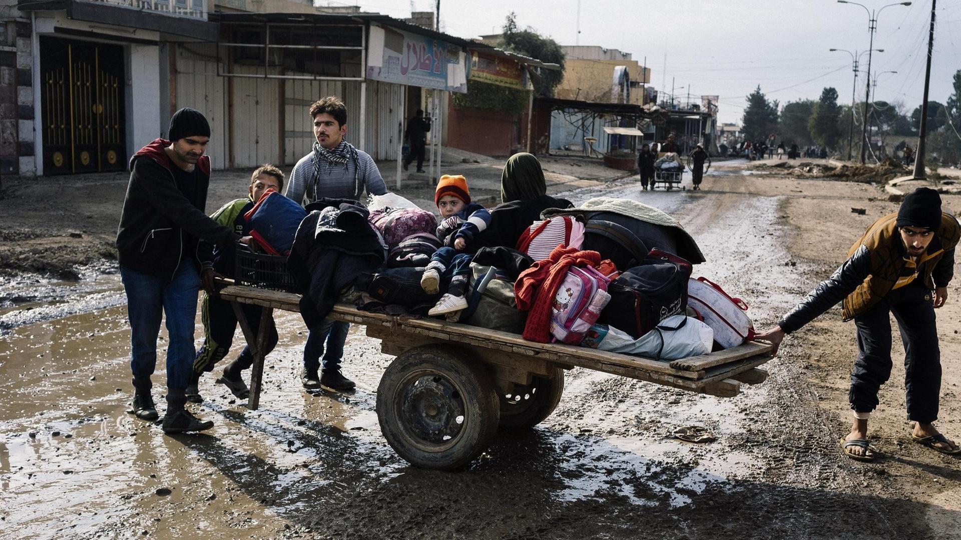 Eine Familie zieht einen improvisierten Wagen mit Kindern und Gepäck über eine schlammige Straße.