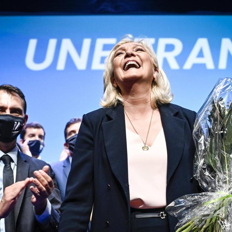 Die Vorsitzende des Rassemblement National, Marine Le Pen, steht auf einer Bühne vor einem Transparent "Une France". Sie hält einen Blumenstrauß und lacht, hinter ihr stehen viele klatschende Menschen, die FFP-Masken tragen. 04/07/2021