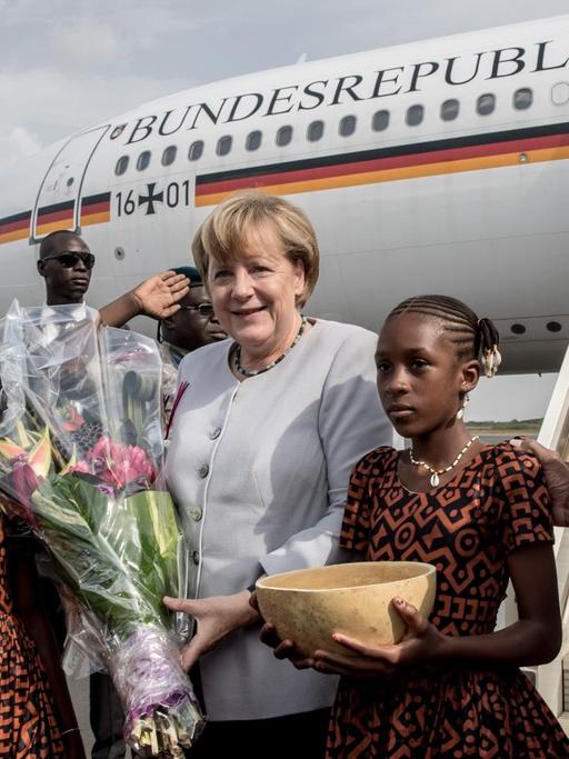 Bundeskanzlerin Angela Merkel steht in Mali vor der Regierungsmaschine. Neben ihr stehen zwei Mädchen mit Blumen und einer traditionellen Wasserkalebasse. Auch Staatspräsident Ibrahim Boubacar Keita ist auf dem Bild zu sehen.