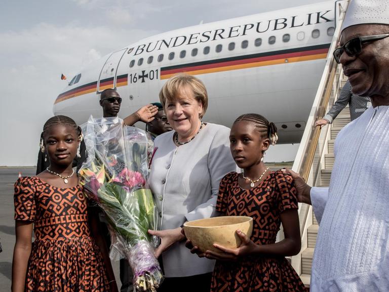 Bundeskanzlerin Angela Merkel steht in Mali vor der Regierungsmaschine. Neben ihr stehen zwei Mädchen mit Blumen und einer traditionellen Wasserkalebasse. Auch Staatspräsident Ibrahim Boubacar Keita ist auf dem Bild zu sehen.