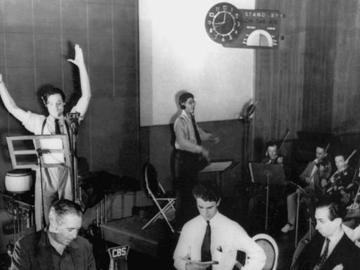 Schwarz-weiß-Aufnahme einer Hörspiel-Übertragung in einem Studio. Sprecher an Mikrofonen und Musiker mit Geigen.
