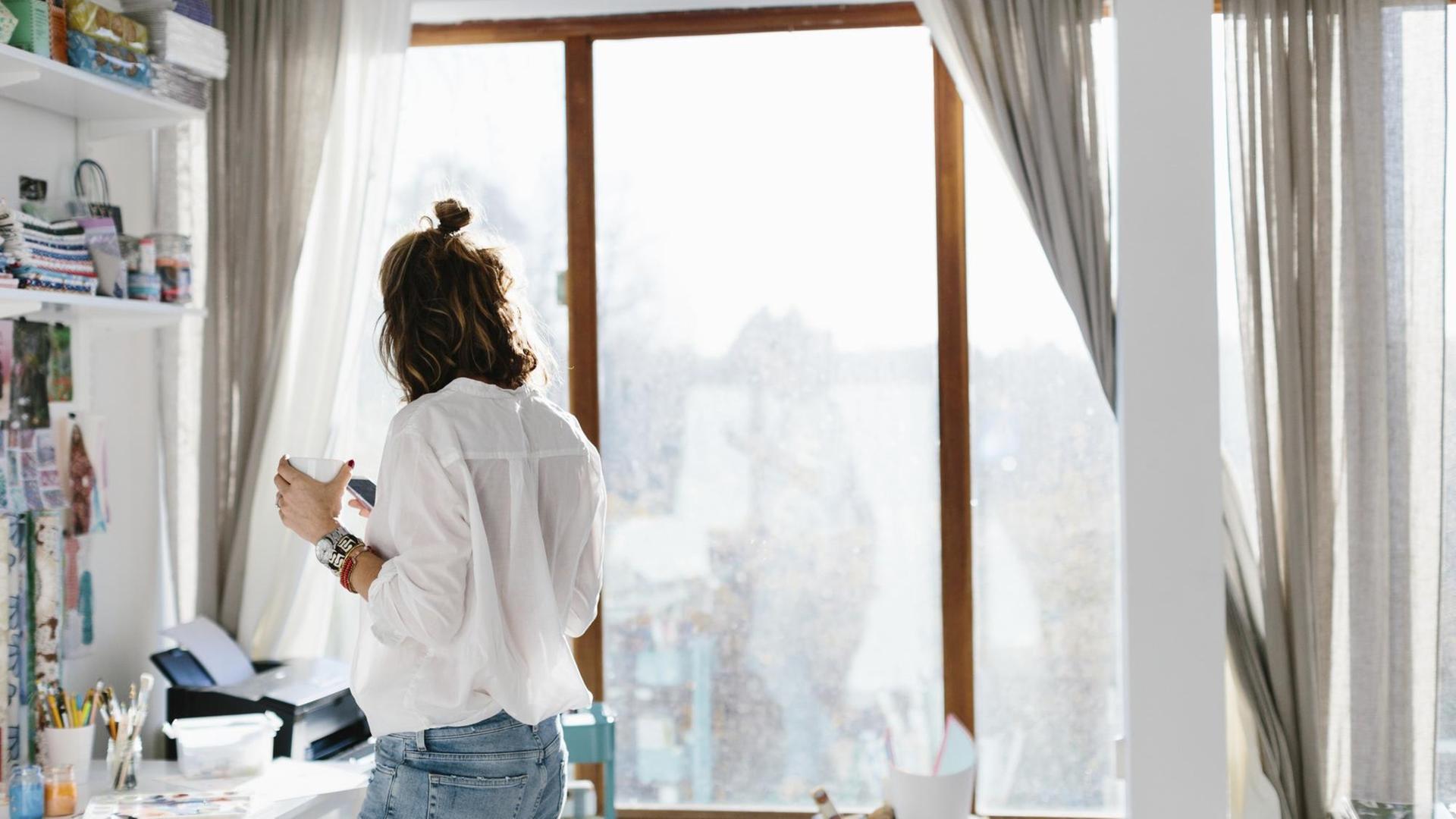 Eine Frau steht mit ihrem Smartphone und einer Tasse Kaffee in der Hand in ihrem Home Office. Eine Frau steht mit ihrem Smartphone und einer Tasse Kaffee in der Hand in ihrem Home Office.