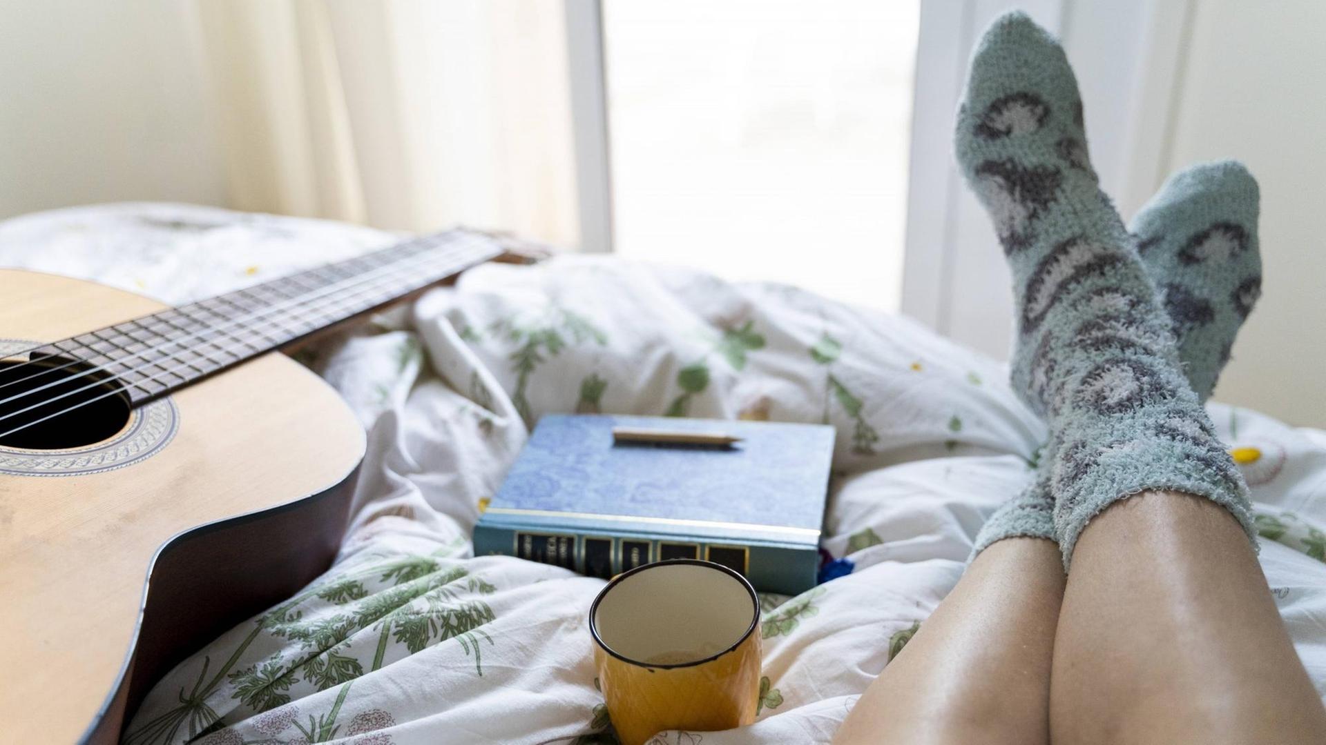 Die Beine einer Frau auf dem Bett, daneben eine Gitarre, ein Buch und eine Tasse. Die Beine einer Frau auf dem Bett, daneben eine Gitarre, ein Buch und eine Tasse.