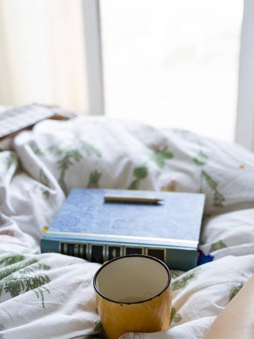 Die Beine einer Frau auf dem Bett, daneben eine Gitarre, ein Buch und eine Tasse.