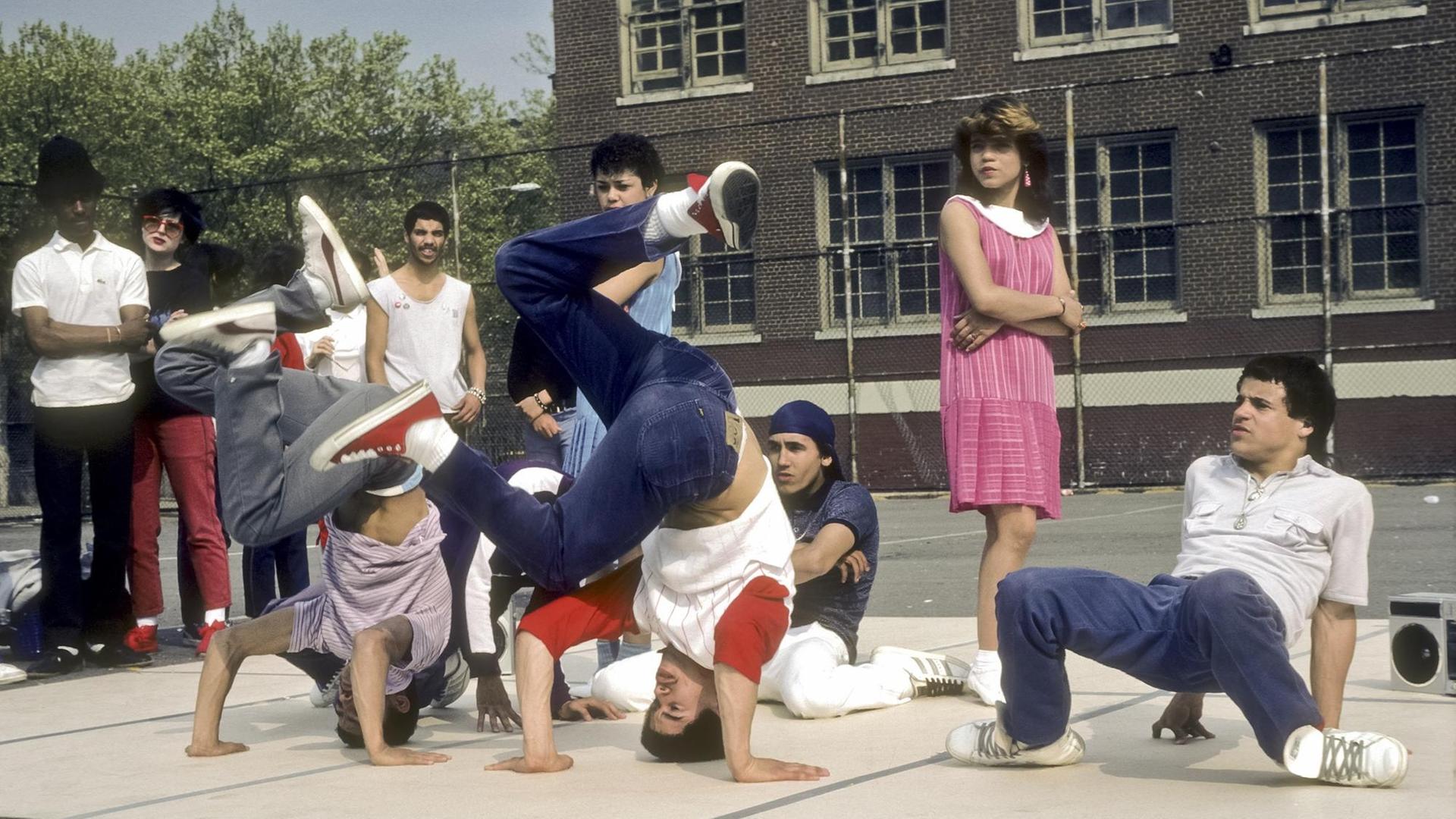 Eine Gruppe Jugendlicher führt auf einem Schulhof eine Breakdance-Choreographie aus, zwei von ihnen befinden sich im Kopfstand und wirbeln mit den Beinen, Umstehende sehen ihnen zu, darunter ein Mädchen in einem langen rosa Kleid. Eine Gruppe Jugendlicher führt auf einem Schulhof eine Breakdance-Choreographie aus, zwei von ihnen befinden sich im Kopfstand und wirbeln mit den Beinen, Umstehende sehen ihnen zu, darunter ein Mädchen in einem langen rosa Kleid.
