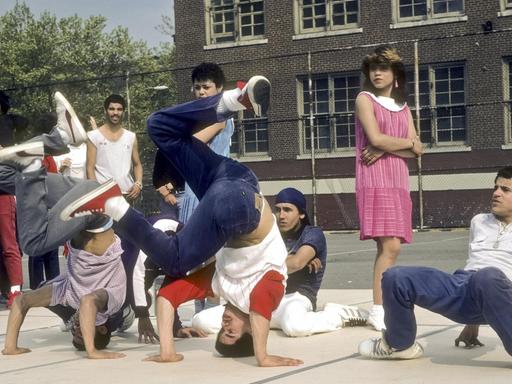 Eine Gruppe Jugendlicher führt auf einem Schulhof eine Breakdance-Choreographie aus, zwei von ihnen befinden sich im Kopfstand und wirbeln mit den Beinen, Umstehende sehen ihnen zu, darunter ein Mädchen in einem langen rosa Kleid.