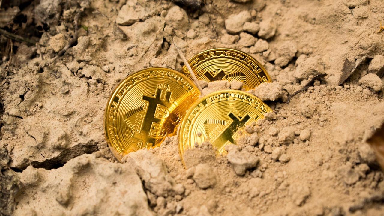 Drei golden schimmernde Bitcoin-Münzen liegen, halb vergraben, im Sand.