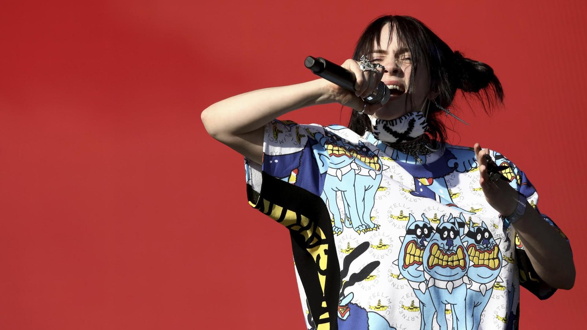 Die Sängerin Billie Eilish steht auf einer Bühne mit rotem Hintergrund, sie trägt ein buntes T-Shirt in der rechten Hand hält sie ein Mikrofon in das sie singt. Die Sängerin Billie Eilish steht auf einer Bühne mit rotem Hintergrund, sie trägt ein buntes T-Shirt in der rechten Hand hält sie ein Mikrofon in das sie singt.
