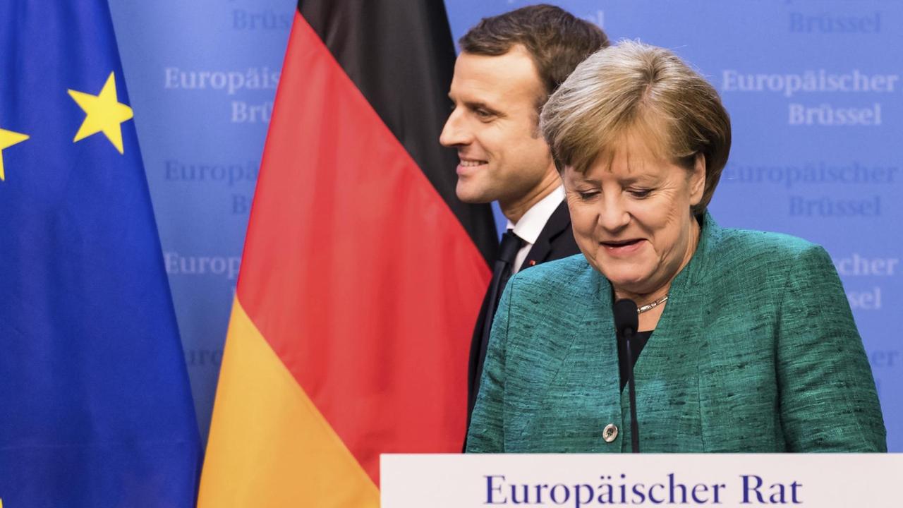 Merkel steht lächelnd am Rednerpult, hinter ihr läuft ebenfalls lächelnd Macron vorbei. An der Wand eine deutsche und eine EU-Flagge. Merkel steht lächelnd am Rednerpult, hinter ihr läuft ebenfalls lächelnd Macron vorbei. An der Wand eine deutsche und eine EU-Flagge.