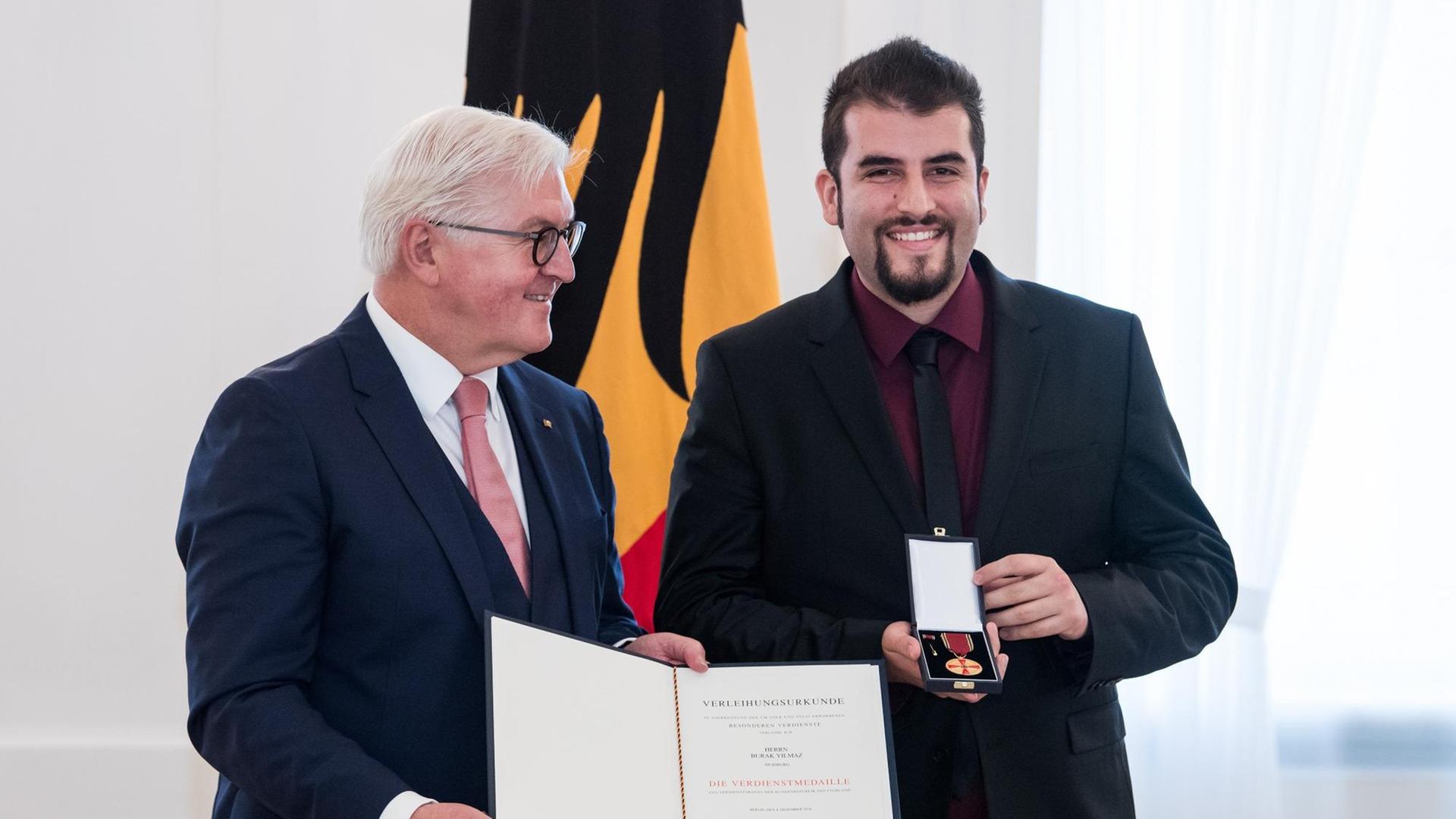 Frank-Walter Steinmeier und Burak Yilmaz stehen nebeneinander in einem festlich geschmückten Raum. Steinmeier mit weißen Haaren hält einen Urkunde in die Kamera, Burak Yilmaz mit schwarzen Haaren und Bart den Verdienstorden der Bundesrepublik Deutschland. Frank-Walter Steinmeier und Burak Yilmaz stehen nebeneinander in einem festlich geschmückten Raum. Steinmeier mit weißen Haaren hält einen Urkunde in die Kamera, Burak Yilmaz mit schwarzen Haaren und Bart den Verdienstorden der Bundesrepublik Deutschland.