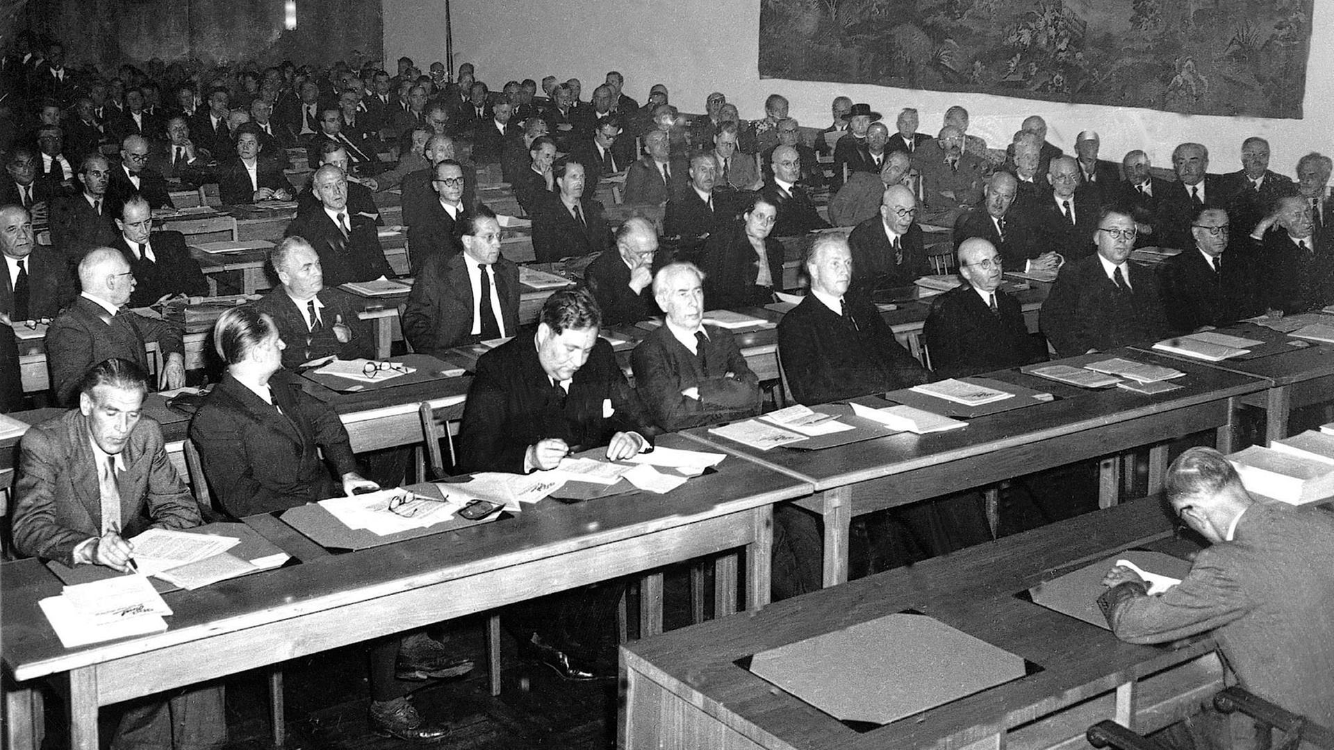 Erste Tagung des Parlamentarischen Rates am Nachmittag des 1. September 1948 in Bonn: Neben den 65 stimmberechtigten Mitgliedern aus westdeutschen Ländern nahmen Vertreter Berlins mit beratender Stimme an der Sitzung teil. In der ersten Reihe von links nach rechts: Max Reimann, KPD; Walter Menzel, SPD; Carlo Schmid, SPD; Theodor Heuss, FDP; Hans-Christoph Seebohm, DP; unbekannt; Adolf Suesterhenn, CDU; Anton Pfeiffer, CDU; Konrad Adenauer, CDU Erste Tagung des Parlamentarischen Rates am Nachmittag des 1. September 1948 in Bonn: Neben den 65 stimmberechtigten Mitgliedern aus westdeutschen Ländern nahmen Vertreter Berlins mit beratender Stimme an der Sitzung teil. In der ersten Reihe von links nach rechts: Max Reimann, KPD; Walter Menzel, SPD; Carlo Schmid, SPD; Theodor Heuss, FDP; Hans-Christoph Seebohm, DP; unbekannt; Adolf Suesterhenn, CDU; Anton Pfeiffer, CDU; Konrad Adenauer, CDU