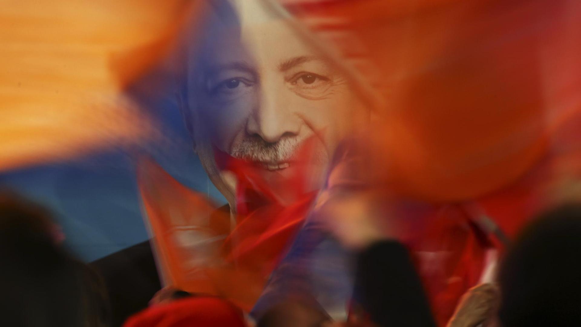 Hinter den unscharf abgebildeten Fahnen und den Köpfen von Menschen zeichnet sich scharf das Bild des lächelnden Erdogan ab. Hinter den unscharf abgebildeten Fahnen und den Köpfen von Menschen zeichnet sich scharf das Bild des lächelnden Erdogan ab.