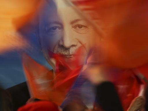 Hinter den unscharf abgebildeten Fahnen und den Köpfen von Menschen zeichnet sich scharf das Bild des lächelnden Erdogan ab.